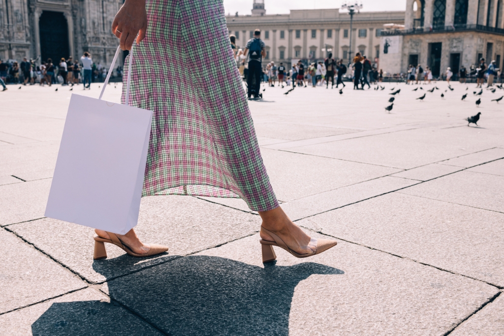 woman in a pleaded pink and green skirt with beige shoes holding a white shopping bag in milan