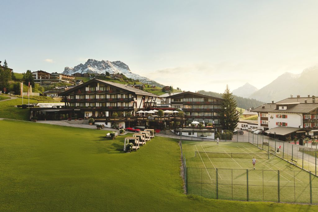 burg hotel facade with alps in the background, green landscaping, and tennis court
