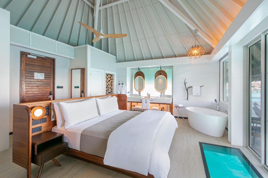 overwater bungalow room with bed, free-standing tub, glass paneled floor, ensuite bathroom, and balcony